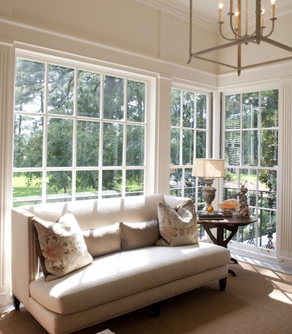 sunroom with large windows overlooking a backyard, a beige sofa with 4 pillows, a wooden side table holds a lamp and two small sculptures 