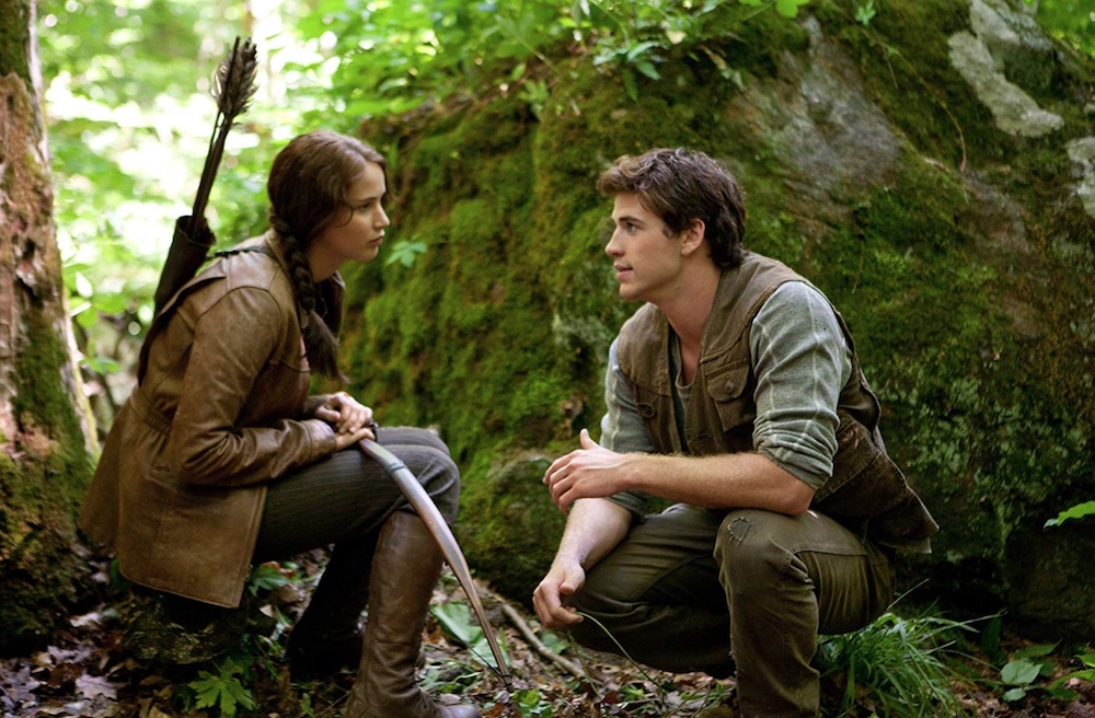 Jennifer Lawrence as Katniss Everdeen on a tree stump in wooded area facing Liam Hemsworth as Gale Hawthorne, leaves and moss all around them