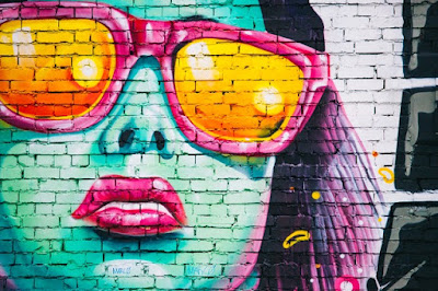 girl painted on wall - street art. Her skin is blue, she is wearing large orange sunglasses, and has purple hair