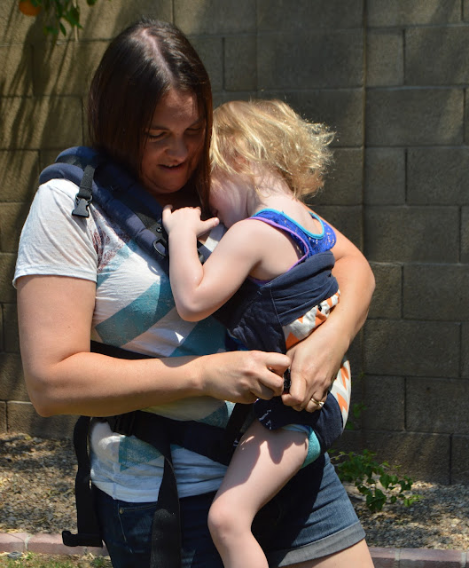 Momma is seen buckling the shoulder strap into the buckle on the carrier, baby is braced by her other arm across the back