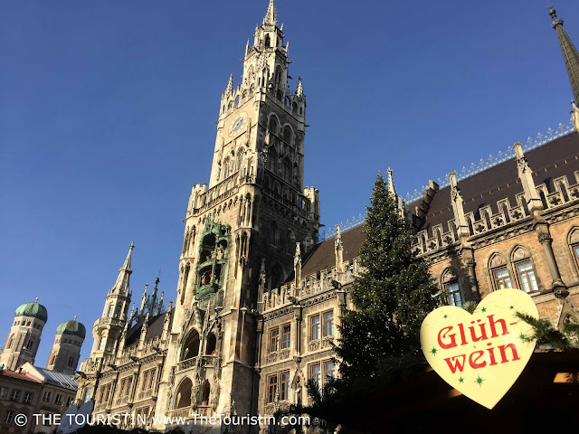 A giant fir tree decorated with Christmas ornaments next to a heart-shaped sign with the word Glühwein in red letters, in front of a tall stone tower under a bright blue sky.