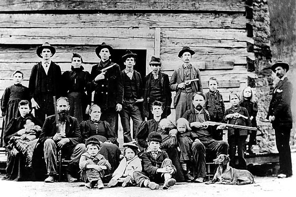 around 20 people (adults and children, plus a dog), all in their "Sunday best," gathered for a black-and-white formal group photo in front of a log cabin