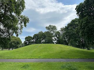 A photo showing grass in the foreground a grey path and then a grassy hillock behind.  At either side is a line of large, old trees.  Photograph by Kevin Nosferatu for the Skulferatu Project.
