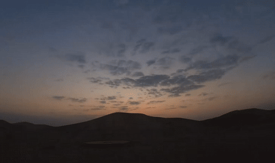 A time lapse of the rising sun slowly illuminating the dunes and distant hills of the Varzaneh Desert, as scattered clouds glow in warm morning light.