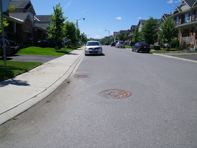 Suburban street with sidewalk adjacent to the roadway with a flat sidewalk and the entire length of the curb is ramped from the sidewalk height to the roadway. (Rosehill at Huntmar Drive, Stittsville)