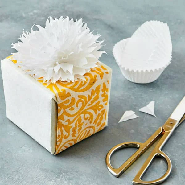 white cupcake liner flower that resembles chrysanthemum on top of wrapped yellow and white cube gift box displayed alongside pair of gold and silver scissors