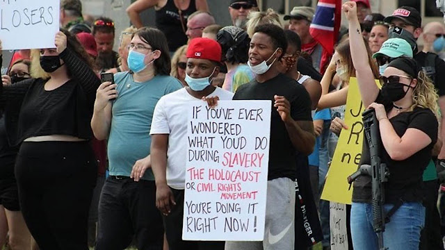 This photo shows a crowd of the original protest group, who want the statue removed. The crowd contains both White and Black people, many of whom are wearing masks. A protester at the front of the crowd holds a sign that reads, “If you’ve ever wondered what you’d do during slavery, the Holocaust, or Civil Rights Movement, you’re doing it right now!”