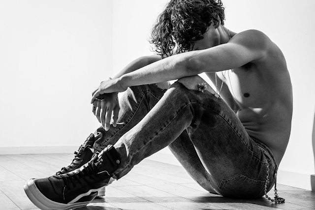 A black and white photo of a shirtless man in jeans crouching by a wall.
