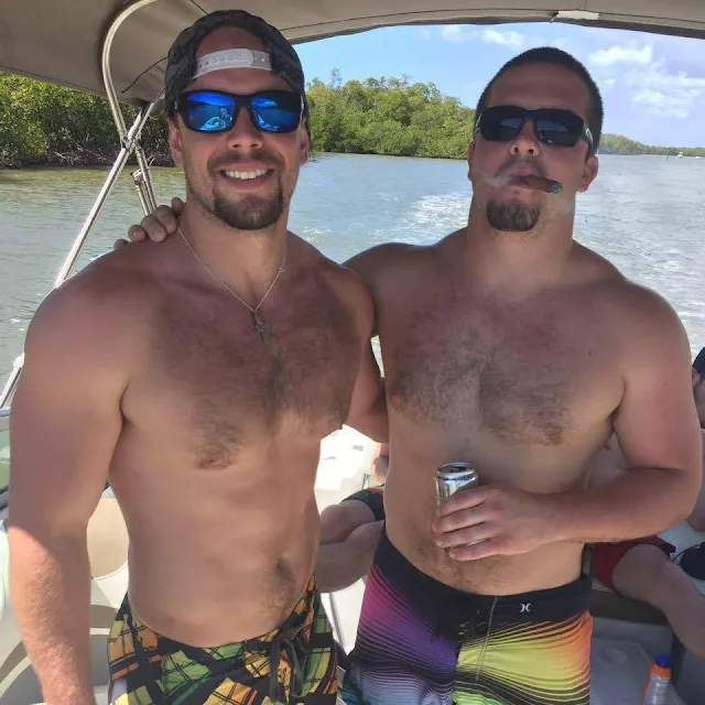 Two very muscular handsome scruffy face sunglasses wearing shirtless men from the waist up smoking cigars and driving a boat