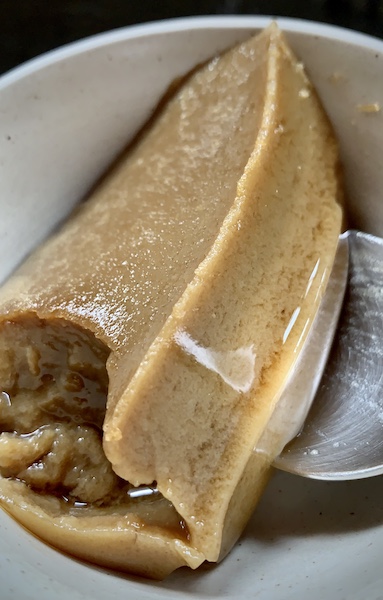 Close up of a sweet called GiNNa in Kannada. This is the vegan, cruelty-free version. Rectangular slice, brown in colour, jelly texture. In an off white bowl and a bit of steel spoon showing.