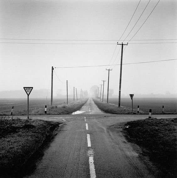A black and white image of a four way crossroads in the middle of nowhere, with a Give Way sign on the left and one on the right facing another direction, and overhead power and telephone wires, in foggy weather. Taken by Paul Hart