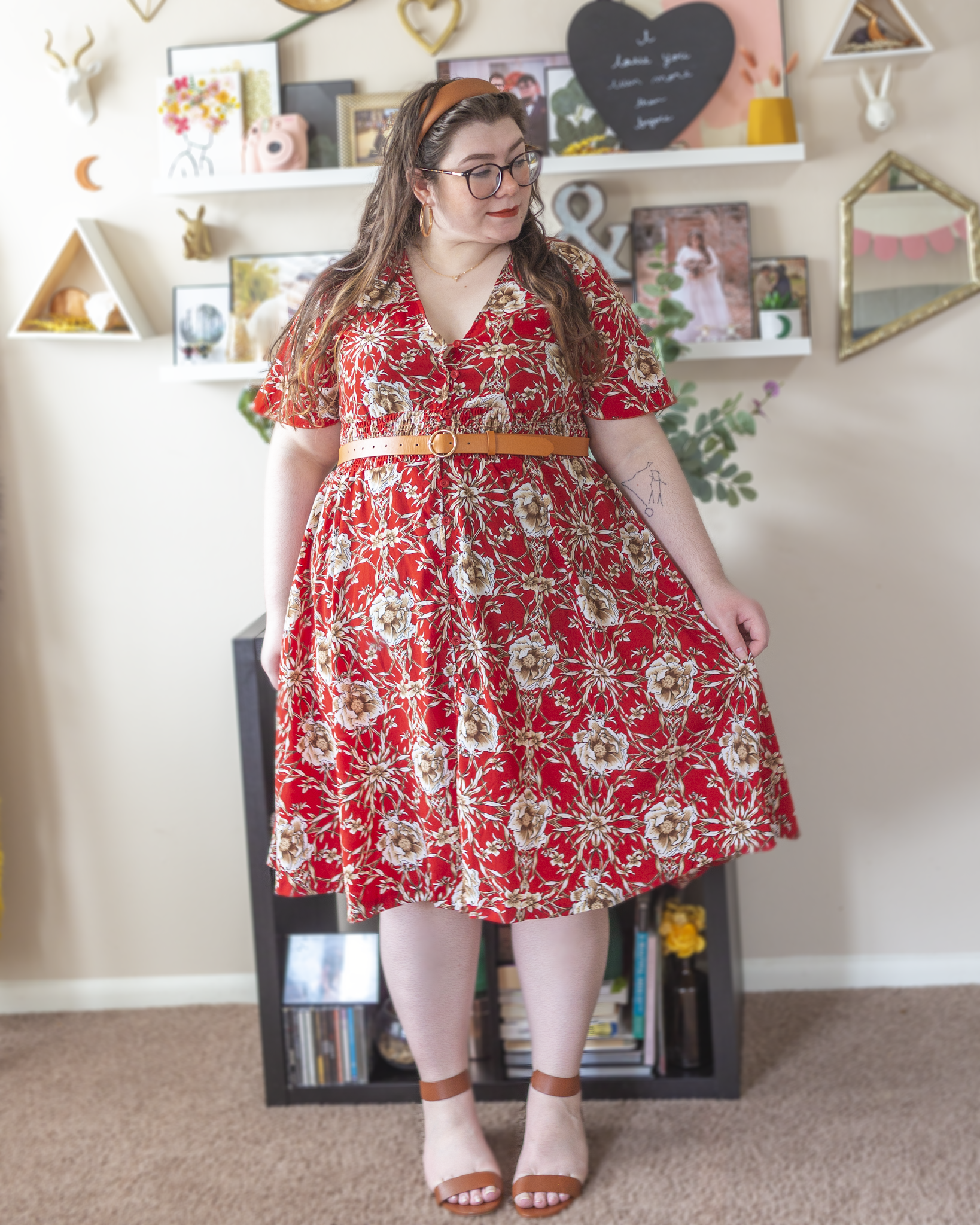 An outfit consisting of a cognac faux leather thick headband, red short flounce sleeve button up with beige floral midi dress, belted with cognac brown belt and a brown heeled ankle strap sandals.