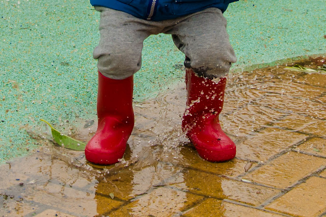 Piernas de mi pequeña con las botas de agua rojas, saltando sobre charco grande de agua. 