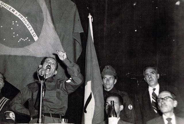 Na foto podemos ver Plínio Salgado, líder do movimento integralista brasileiro, discursando em um palanque com a bandeira brasileira ao fundo.