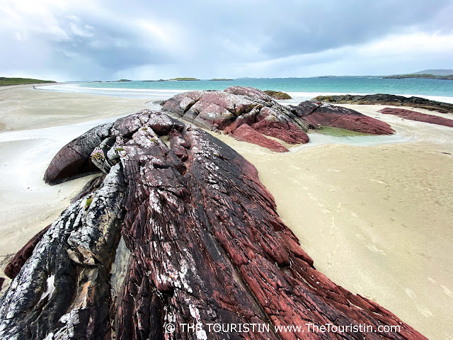 Red-coloured rock formations on a white sand beach and the green-blue-coloured ocean under a cloudy sky.