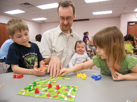 A man sits at a table with two children who appear to be in kindergarten or first grade, playing a board game with them. The game is a modular board made up of tiles containing roads through grassy fields, and the players are placing brightly coloured plastic pieces on the board.