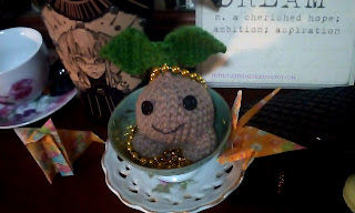 A close shot of several items on my dresser.  The picture is centered on a crocheted mandrake sprouting plushie sitting inside of a teacup filled with gold colored mardi gra beads.  To left side of the tea cup is a leather bracer with a tiefling girl painted onto it and another teacup.  To the right and in the background is a woodensign that says, "Dreams n. a cherished hope; ambition; aspiration."  Though the word Dream is only partly in the frame.  There are also two origami animals on the surface of the dresser.  