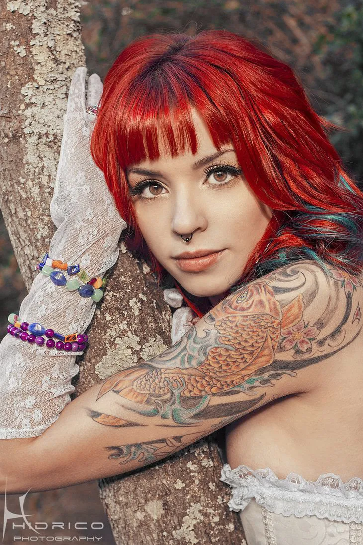Vemos a chica pelirroja apoyada en arbol, está vestida de novia con tatuaje de carpa en el brazo