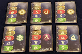 Six action cards laid out side-by-side. Each card has five symbols on the left half, each symbol in a different coloured space. The top space on each card is white; the space below that is yellow, the middle space is red, then a blue space, and a green space at the bottom. The right half of each card has one of five different icons: a CPU on a yellow circle, a horseshoe magnet on a red circle, an atom symbol on a green circle, a chemistry vial on a purple circle, or a microscope on a blue circle.