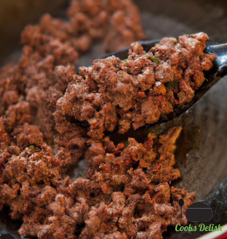 A close-up image of the seasoned ground beef cooking in a skillet. The beef is sizzling and the spices are releasing an aromatic aroma. The beef is being stirred and broken up into small pieces with a spatula.