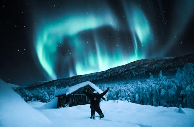 Person standing in front of a cottage with aurora borealis on the horizon