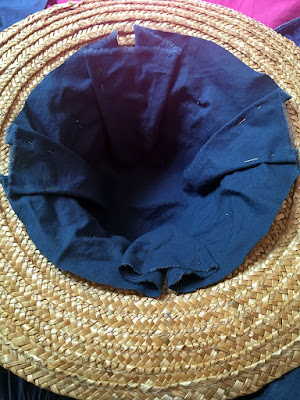 An upside-down straw hat, with medium-blue cotton fabric pushed into the crown, pleated at four points to fit more smoothly, and pinned with silver pins. The edges of the fabric have been roughly trimmed to stop just past the transition from crown to brim.