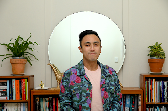 Chinese man in coulourful jacket standing in front of a large round mirror.