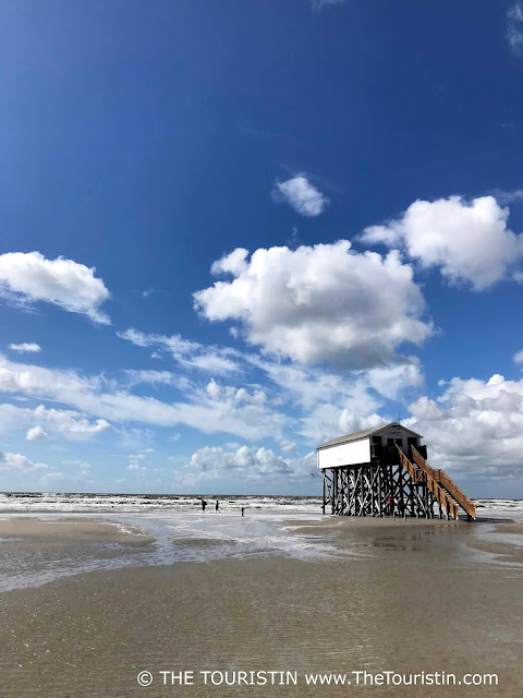 Three people at low tide next to a white wooden cottage on stilts in the sea under a bright blue sky dotted with clouds.