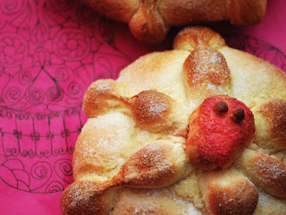 A closeup of a Pan de Muerto (Bread of the Dead) roll. It is decorated with a red skull on top and sits on a pink cloth.