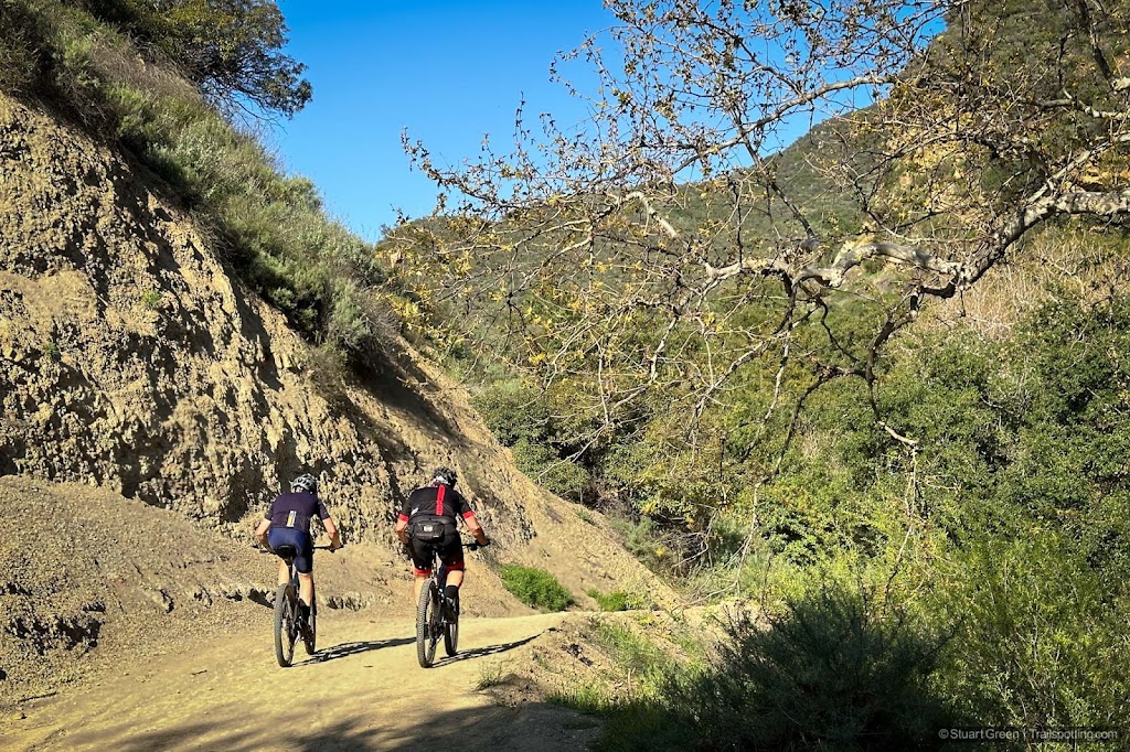 Two mountain bikers on a dusty access trail. Mountains in the distance.
