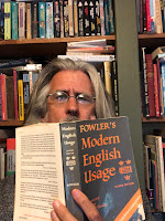 A long haired man peers over the book "Fowler's Modern English Usage." Bookshelf in background.