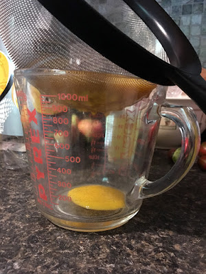 A clear pyrex beaker with red markings and a tiny pool of mango juice on the bottom, with a large black-handled strainer full of pureed mango balanced on top. A few droplets of mango are hanging from the lowest curve of the strainer.