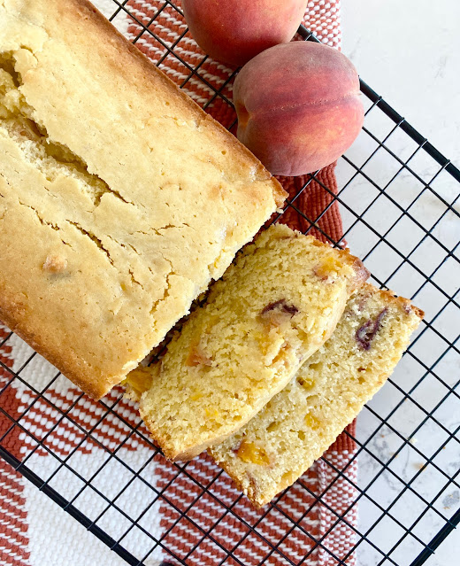 Loaf of peach bread on a wire cooking rack, sliced next to fresh peaches.