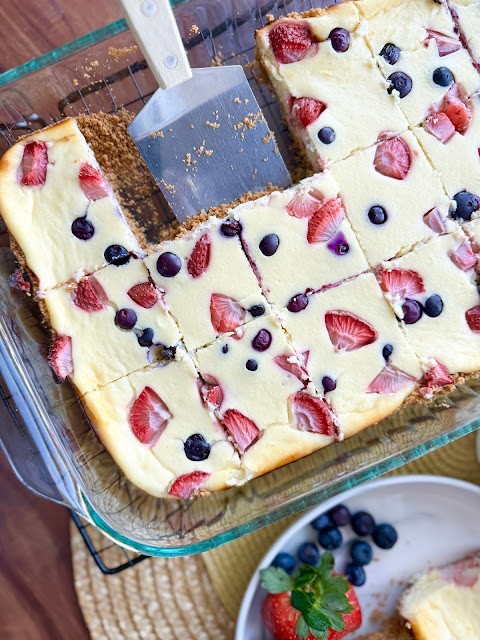 A tray of cottage cheese berry cheesecake bars in a glass baking dish, with a spatula, strawberries and blueberries.