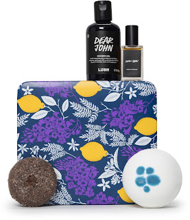 A big blue rectangular box with a circular blue purple label with Dear John in blue with a circular purple shampoo bar with brown circular coffee beans embedded, a cylindrical bottle with purple liquid with a black label saying Dear John shower gel in white font, a spherical white bath bomb with blue spot on the top and a rectangular glass bottle of the perfume on a bright background 