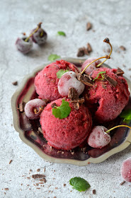 Drei Kugeln Kirschensorbet auf einem kleinen silbernen Teller, garniert mit Schokostückchen, gefrorenen Kirschen und grünen Blättchen.