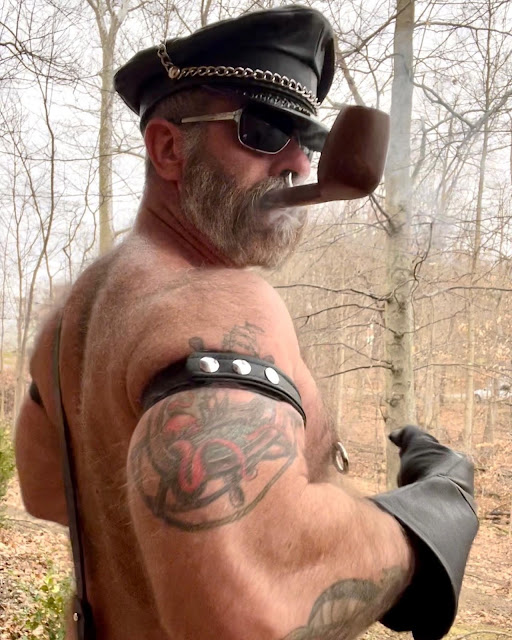 Standing in the woods shirtless with sunglasses on with a pipe in his mouth wearing leather gloves and arm cuff making muscles