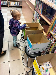 Sala biblioteczna. Dziewczyna w fioletowej wzorzystej bluzie wrzuca do lewego pudełka oznaczonego na zielono kartonik z ilustracją, obok stoi pudełko kartonowe oznaczone na niebiesko – papier i  na żółto – plastik.