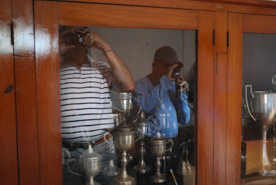 Reflexo de Ramão e Luiz no armário de troféus. O armário é marrom e as portas de vidro, dentro há vários troféus.