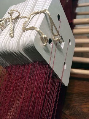 A close-up of neatly arranged white tablet-weaving cards, suspended on strands of extremely fine deep garnet thread thorugh each of four holes and tied with thick shiny white cord at the tops.