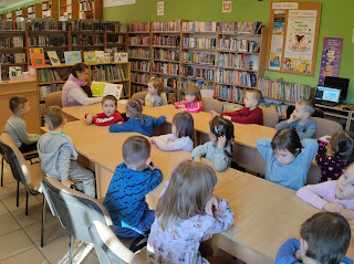 Sala biblioteczna. Pani bibliotekarka siedzi przed grupą przedszkolaków i pokazuje im ilustrację z książki przedstawiająca motyla. Za panią na półregale stoi wystawka książek o motylach i plakat z napisem 24 marca – Dzień Motyla. W tle widzimy regały z książkami, na ścianie wisi tablica korkowa z plakatami promującymi akcję Mała Książka – Wielki Człowiek.