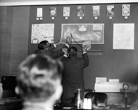 A black and white photograph of two men holding a dowsing rod up against a map on a wall.