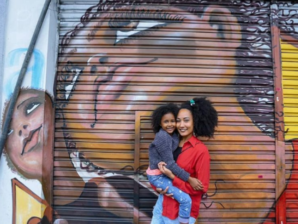 Uma mãe segura a filha no colo diante de um grafite com pessoas negras no desenho. Elas também são negras e sorriem para a câmera.