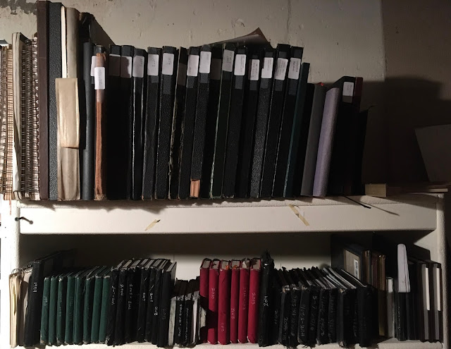 Bookshelf containing numerous sketchbooks of various sizes and colors, many with date labels on spines."