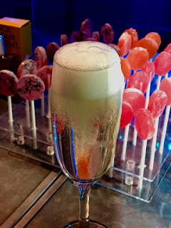 A long thin champagne flute filled with clear prosecco topped with a thick layer of white foam with a wooden cylindrical thin stick poking out of the top on a rectangular silver table with a blue cloth on a dark background 