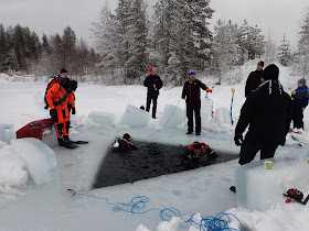 Kaksi sukeltajaa kolmionmallisessa avannossa, ympärillä jäällä muita ihmisiä ja kolmas sukeltaja