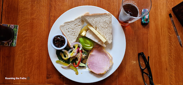 White place on a Wood table with Bread, Butter, Cheddar, Brie, Salad of leaves and mixed peppers, Ham andpickle.