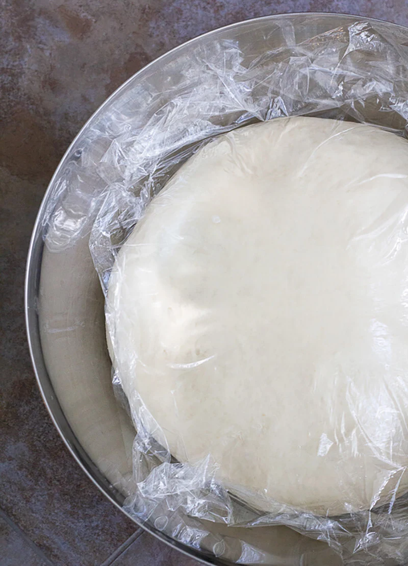 Butter bread mixture in a silver covered with plastic wrap as it yields.