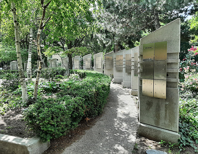 A number of standing pillars arranged in an arc within a park. Each pillar has 6 stainless plaques affixed to it, with names engraved on each.