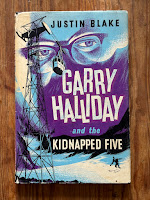 Cover of Garry Halliday and the Kidnapped Five by Justin Blake (Faber, 1962) with artwork by Leo Newman in black, white, blue and purple showing silhouette of skier on snow below a cable car, with close up of eyes behind glasses in background.